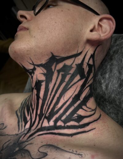 A close-up of a custom freehand abstract blackwork lettering tattoo covering a client's neck and upper chest, showcasing neotribal and dark-themed calligraphy elements by London tattoo artist Goorazz at Parliament Tattoo