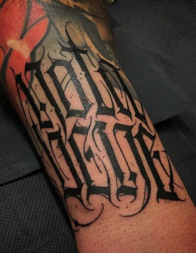 A client's forearm displaying a bold, custom freehand blackwork tattoo by Goorazz, featuring gothic calligraphy lettering with abstract flourishes and a dark theme, created in London