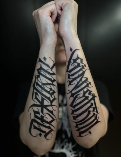 A close-up of a person's forearms showcasing two large, bold freehand gothic lettering tattoos in a custom blackwork script style by tattoo artist Goorazz