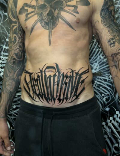 A client's stomach featuring a large, custom freehand blackwork tattoo by Goorazz, showcasing bold dark-themed gothic calligraphy and abstract lettering elements, created in London at Parliament Tattoo