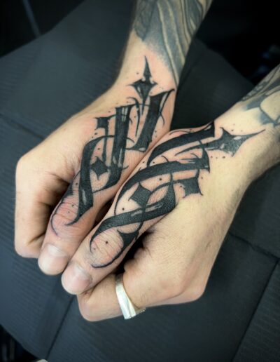 A close-up of a client's hands showcasing bold, custom freehand blackwork finger thumbs tattoos by Goorazz, blending gothic calligraphy with abstract and dark-themed artwork, created in London.
