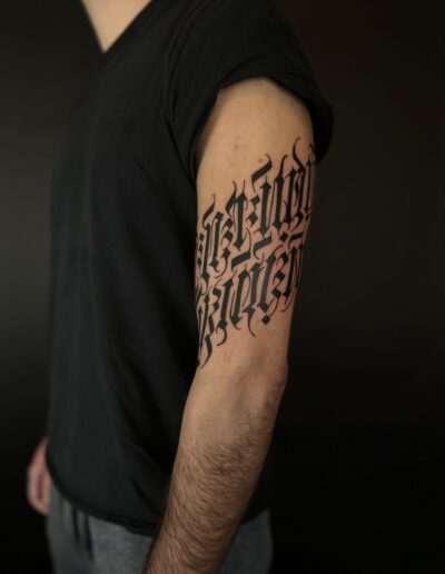 A client's bicep featuring a large, bold custom freehand gothic calligraphy lettering tattoo in blackwork by Goorazz, a London artist specializing in dark-themed designs.