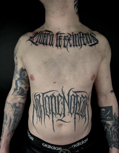 A client's torso featuring two large custom freehand blackwork tattoos by Goorazz, displaying bold dark-themed gothic and abstract calligraphy lettering on the chest and stomach, created in London at Parliament Tattoo