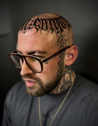 A close-up of a client's shaved head featuring a custom freehand blackwork tattoo by Goorazz at Parliament Tattoo in London, blending bold gothic lettering with abstract calligraphy and dark-themed elements.