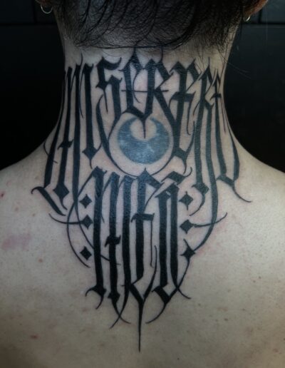 A client's back of the neck featuring a large, custom freehand blackwork tattoo by Goorazz, combining bold gothic calligraphy lettering with abstract elements and a central stylized dark-themed motif.