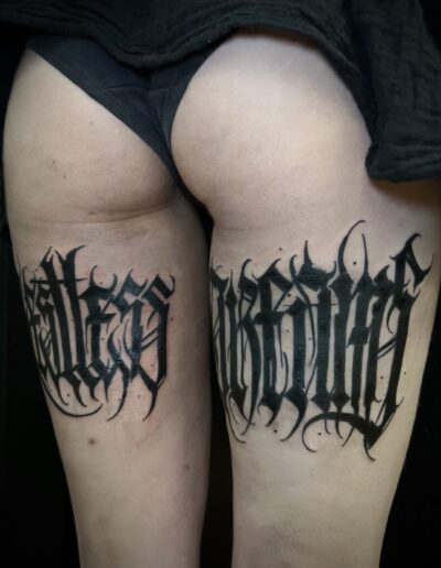 A pair of upper thighs displaying large, custom freehand gothic calligraphy lettering tattoos by Goorazz, featuring dark-themed blackwork and abstract elements, created in London at Parliament Tattoo